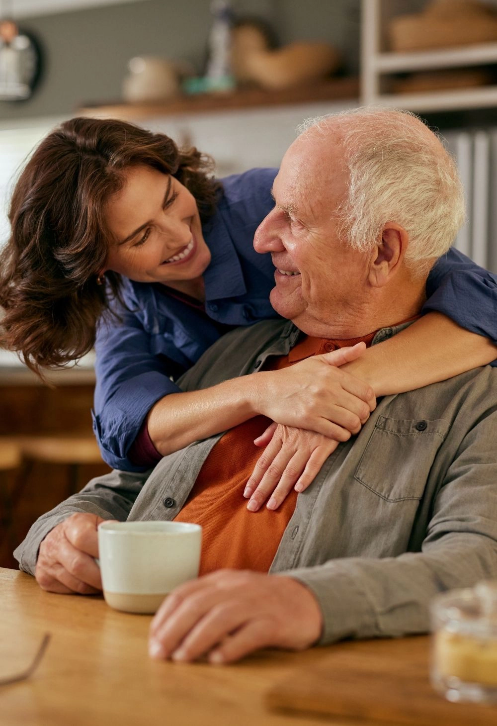 Caregiver affectionately hugging an older man while he enjoys a cup of coffee at home.