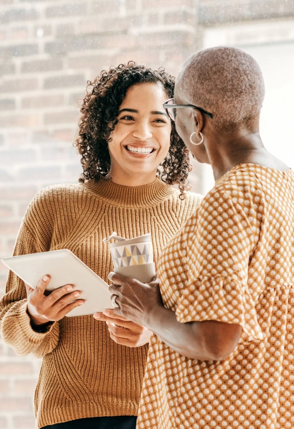 A smiling caregiver assists an older woman by holding a tablet and cup of coffee while they chat together.