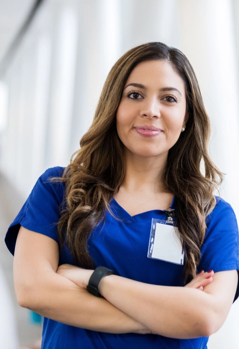 Confident caregiver in blue scrubs standing with arms crossed in a bright hallway.
