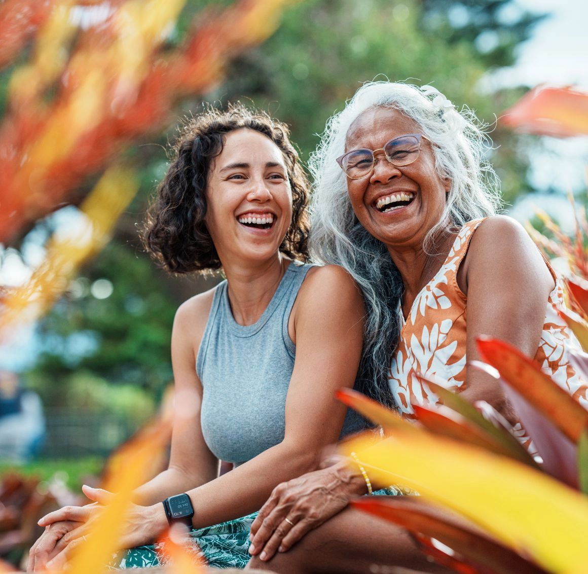 Joyful Companionship Outdoors An older woman and her caregiver share a warm, joyful laugh together while sitting outside among colorful plants.