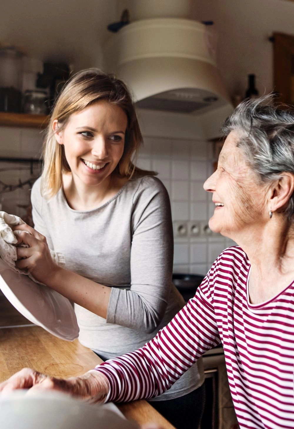 A caregiver helps an elderly woman wash dishes in the kitchen while they smile and talk together.