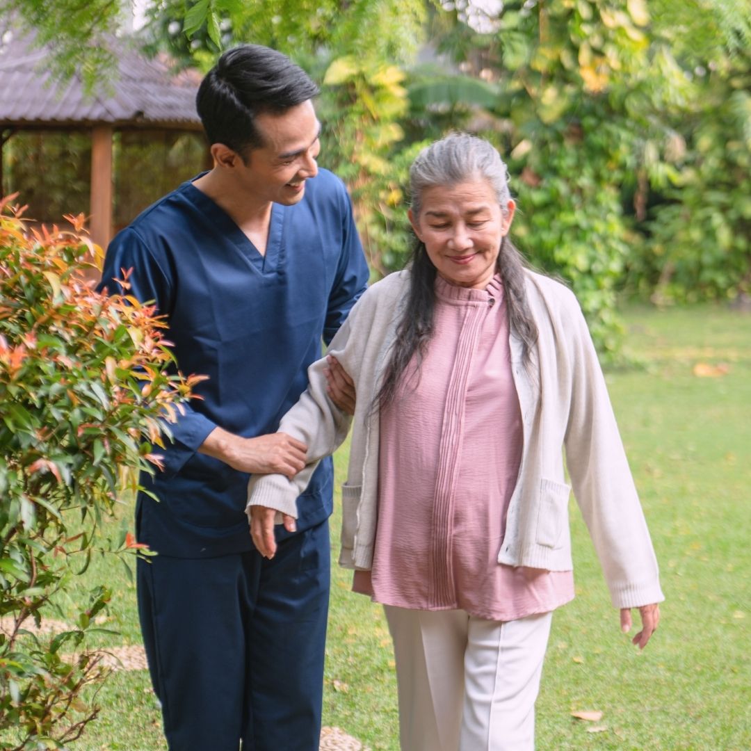 Male nurse assisting an older woman while walking in a garden, showing safe mobility support and home health care in Imperial Valley.