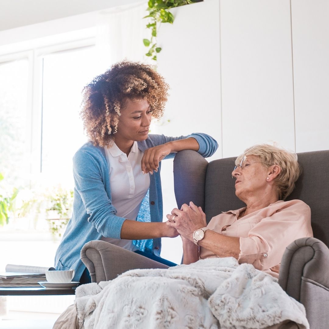 Caregiver supporting an elderly woman resting in a recliner, showing gentle personal care and home assistance services.