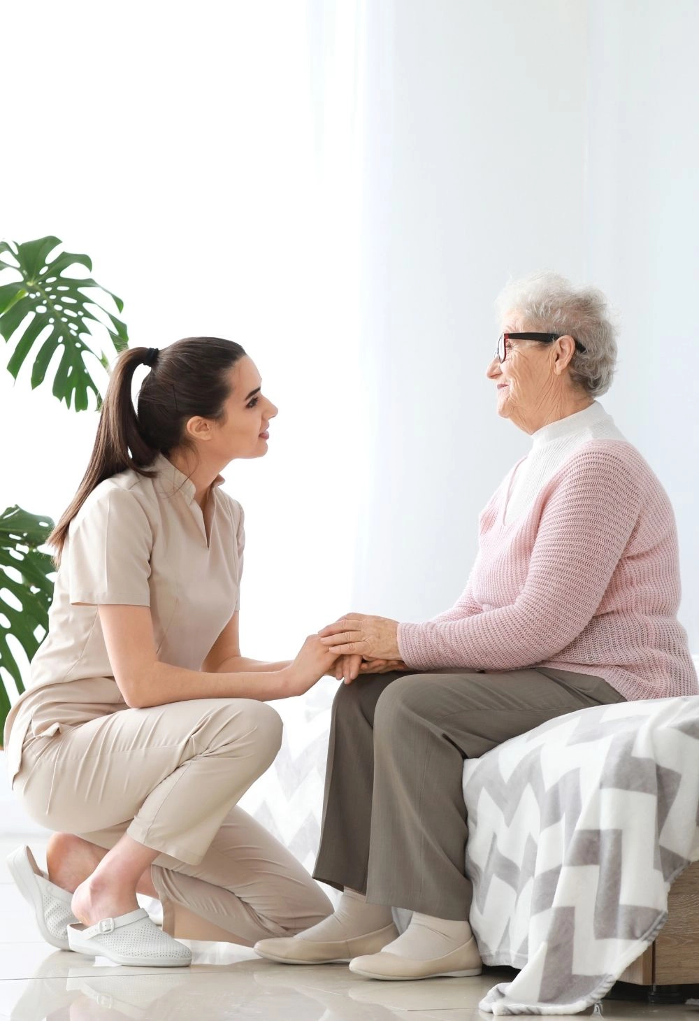 Caregiver kneeling beside an elderly woman while holding her hands and engaging in supportive conversation.
