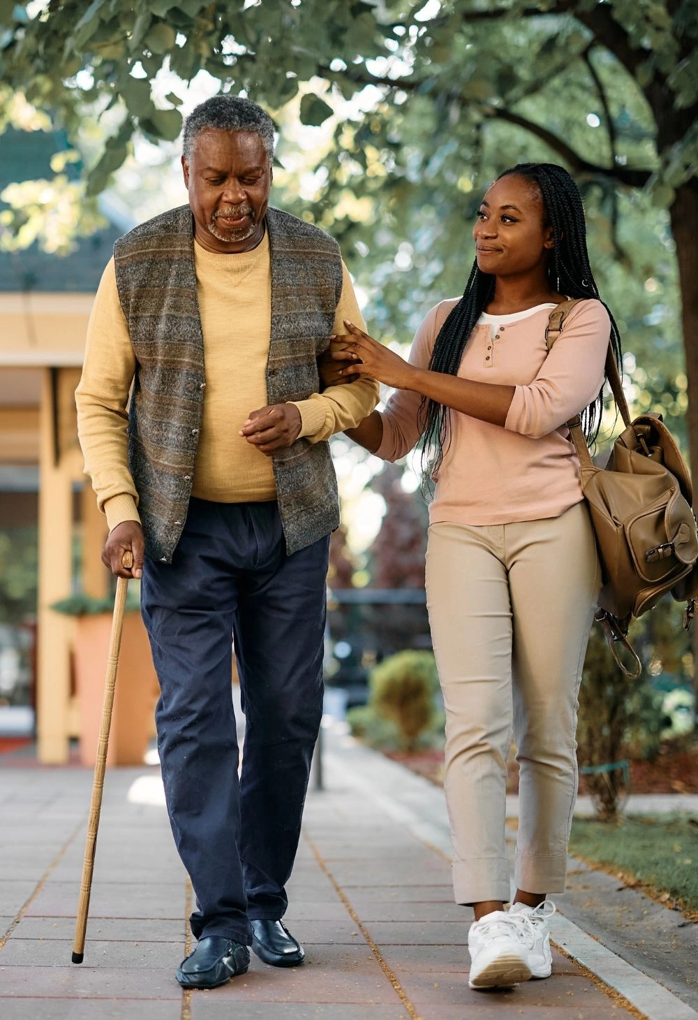 Caregiver supporting an elderly man with a cane while walking outdoors in a peaceful neighborhood.