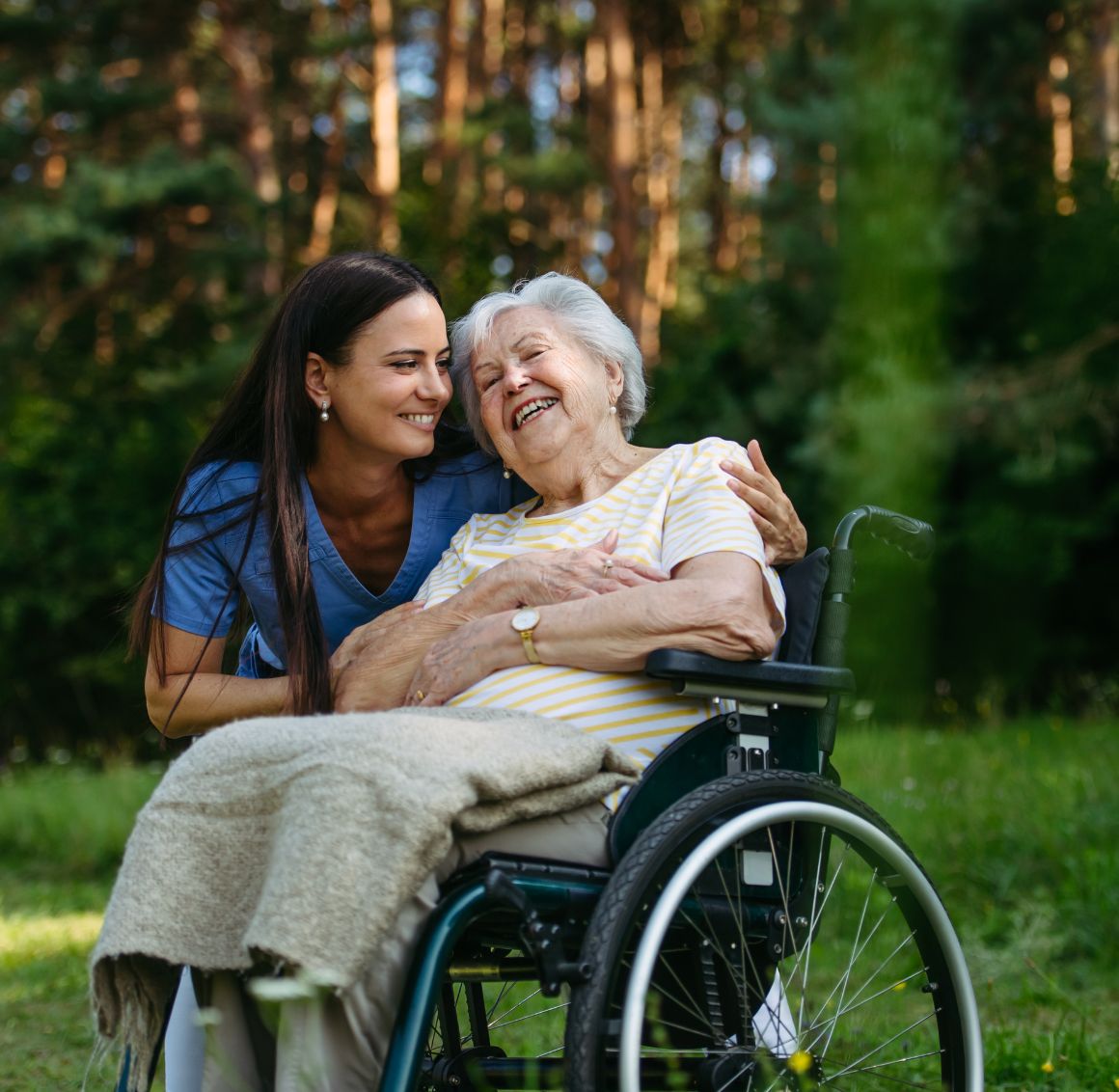 Caregiver smiling and embracing an elderly woman in a wheelchair outdoors, representing compassionate home care in Long Beach, CA.